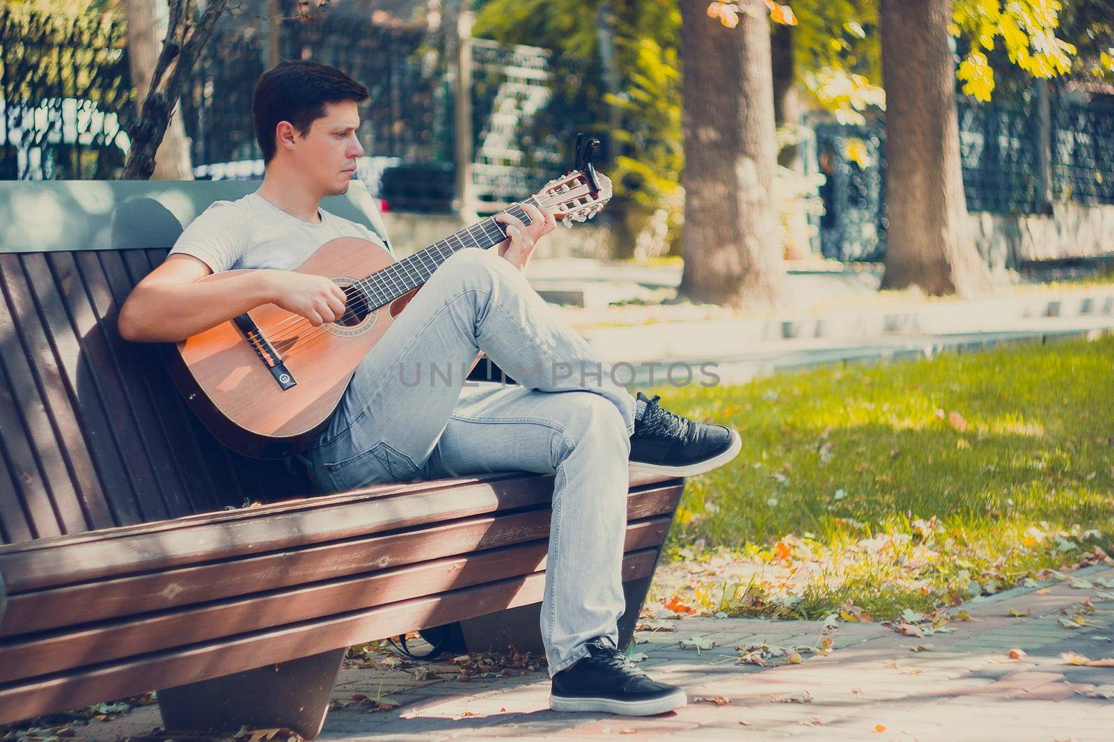 Young man sitting on the bench in the park playing on acoustic guitar with capo. Young attractive man enjoys live music in last sunny days autumn holiday. Retro lens used. Selective soft focus.