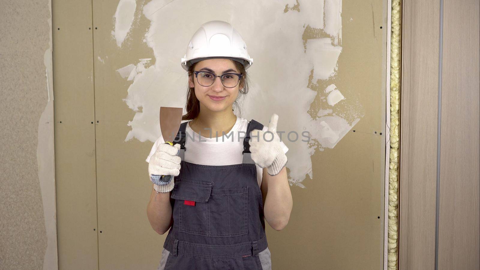 A builder with a spatula in her hand looks at the camera and shows the class and smiles. A woman in special clothes and a helmet makes repairs. 4k