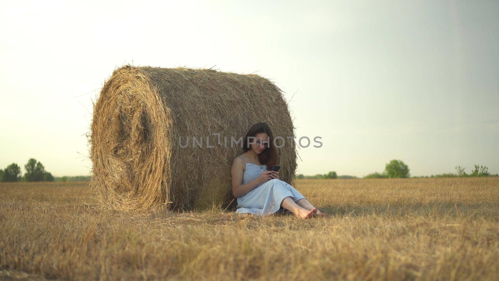 A young woman sits near a haystack in a field with a phone in her hands. A girl with glasses and a blue dress is texting on the phone. 4k