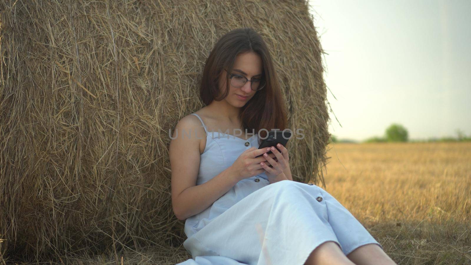 A young woman sits near a haystack in a field with a phone in her hands. A girl with glasses and a blue dress is texting on the phone. 4k
