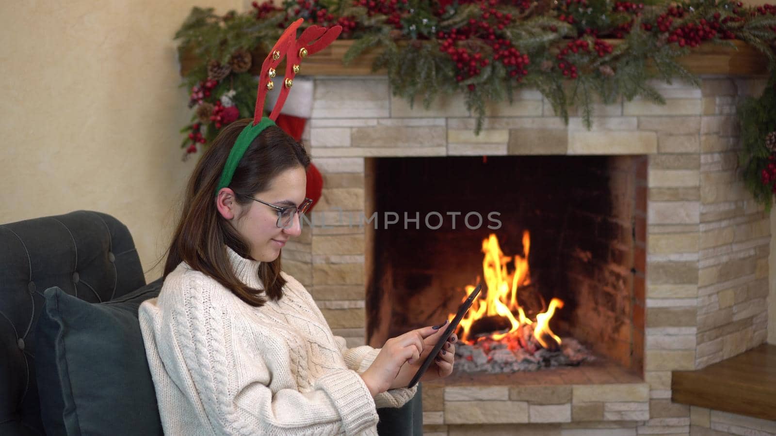 A young pregnant woman with glasses sits in a chair by the fireplace with horns on her head and writes on the tablet. 4k