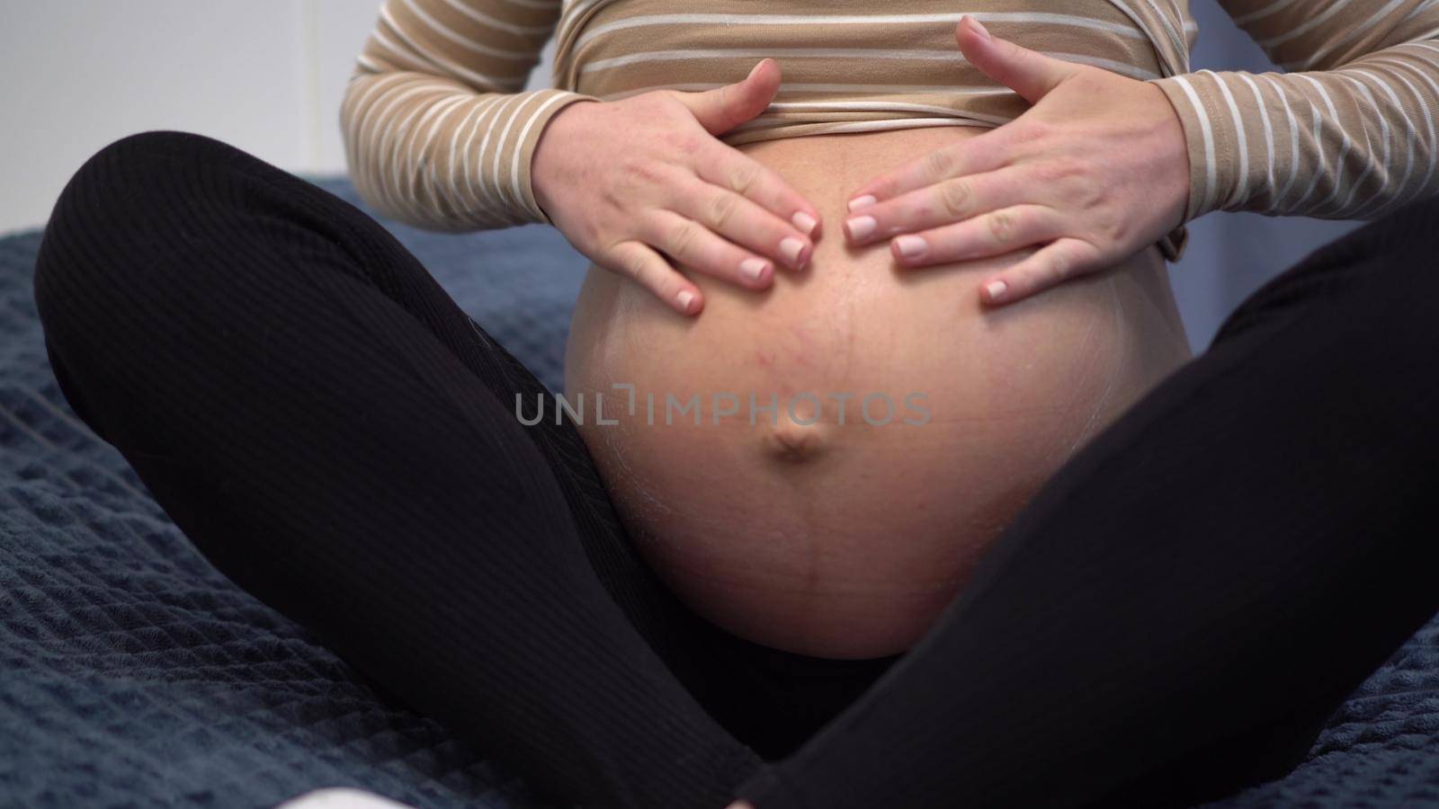 Young pregnant woman with glasses smears cream on her belly. Girl in the bedroom on the bed closeup. 4k
