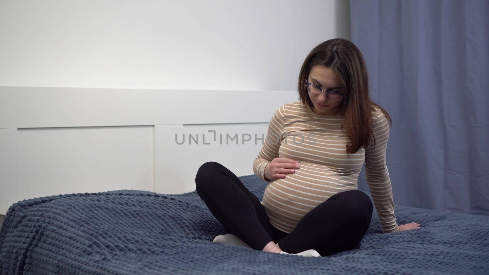 Young pregnant woman with glasses stroking her belly. The girl in the bedroom on the bed. by Puzankov