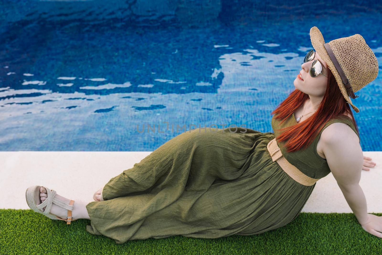 young woman, lying in the sun in front of the pool at home on her summer vacation. girl disconnecting from work. relax and travel concept. Outside, dim sunlight, grass floor with white tiles. copy space.