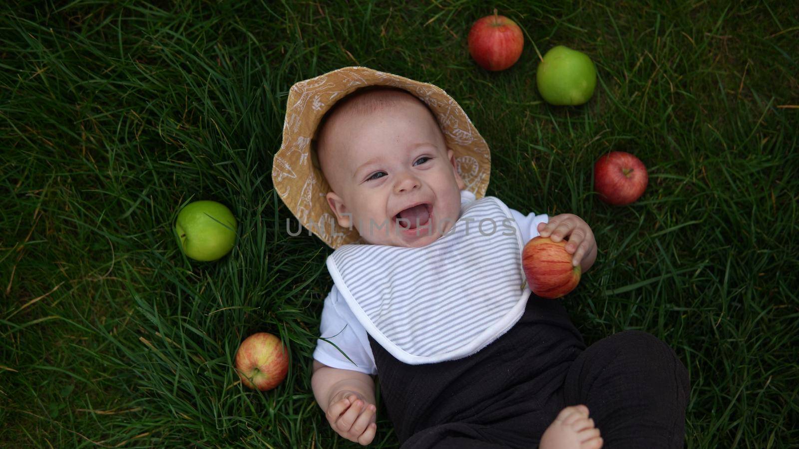 Happy Newborn child in summer panama hat Fall down laying on grass barefoot in Summer Sunny Day with Fresh Fruits Apples. Infant Kid Toddler Boy Smilling Face in Garden Healthy Food Nature harvest.