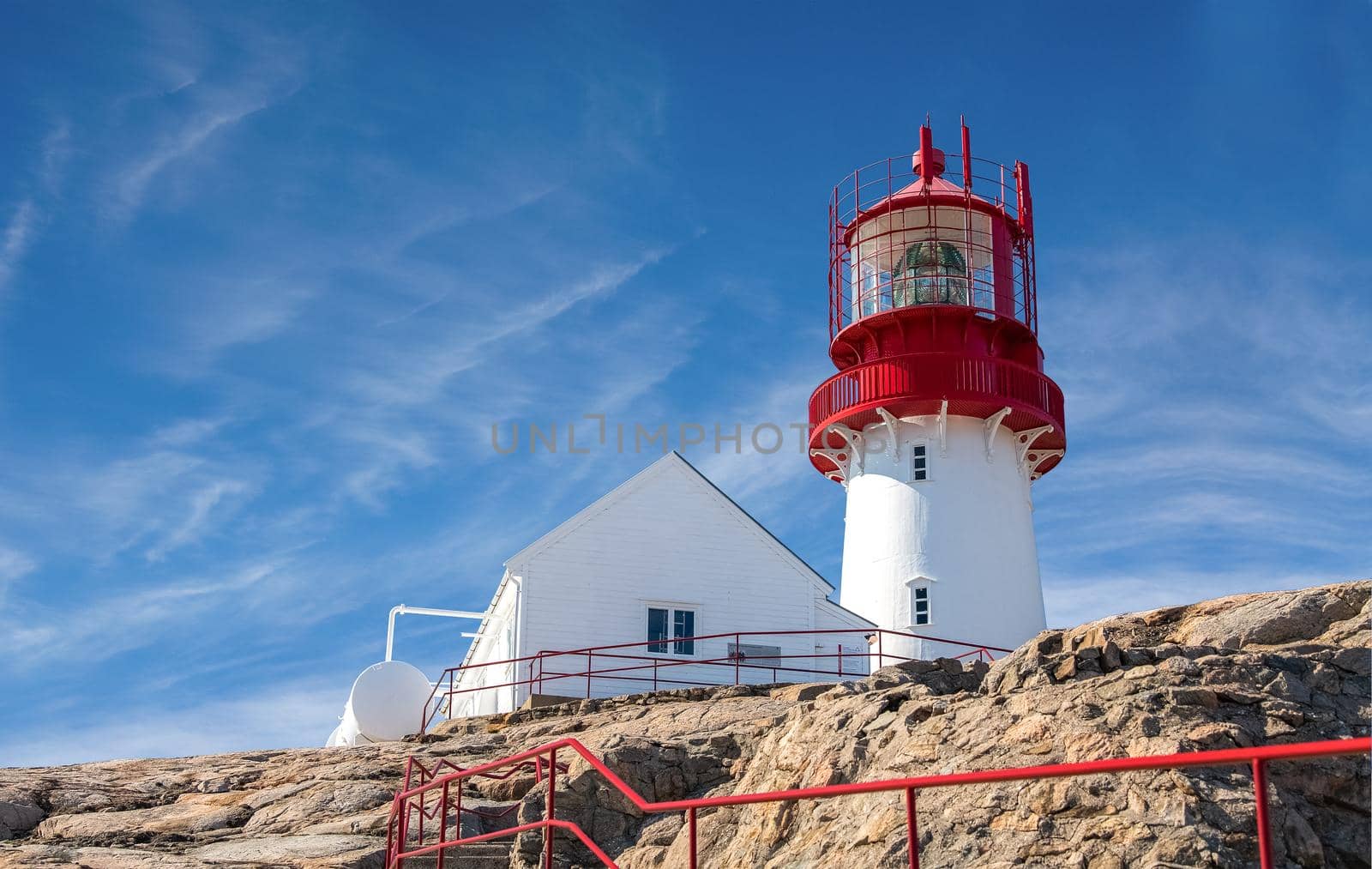 A red lighthouse near a white house on the rocks in the blue ocean