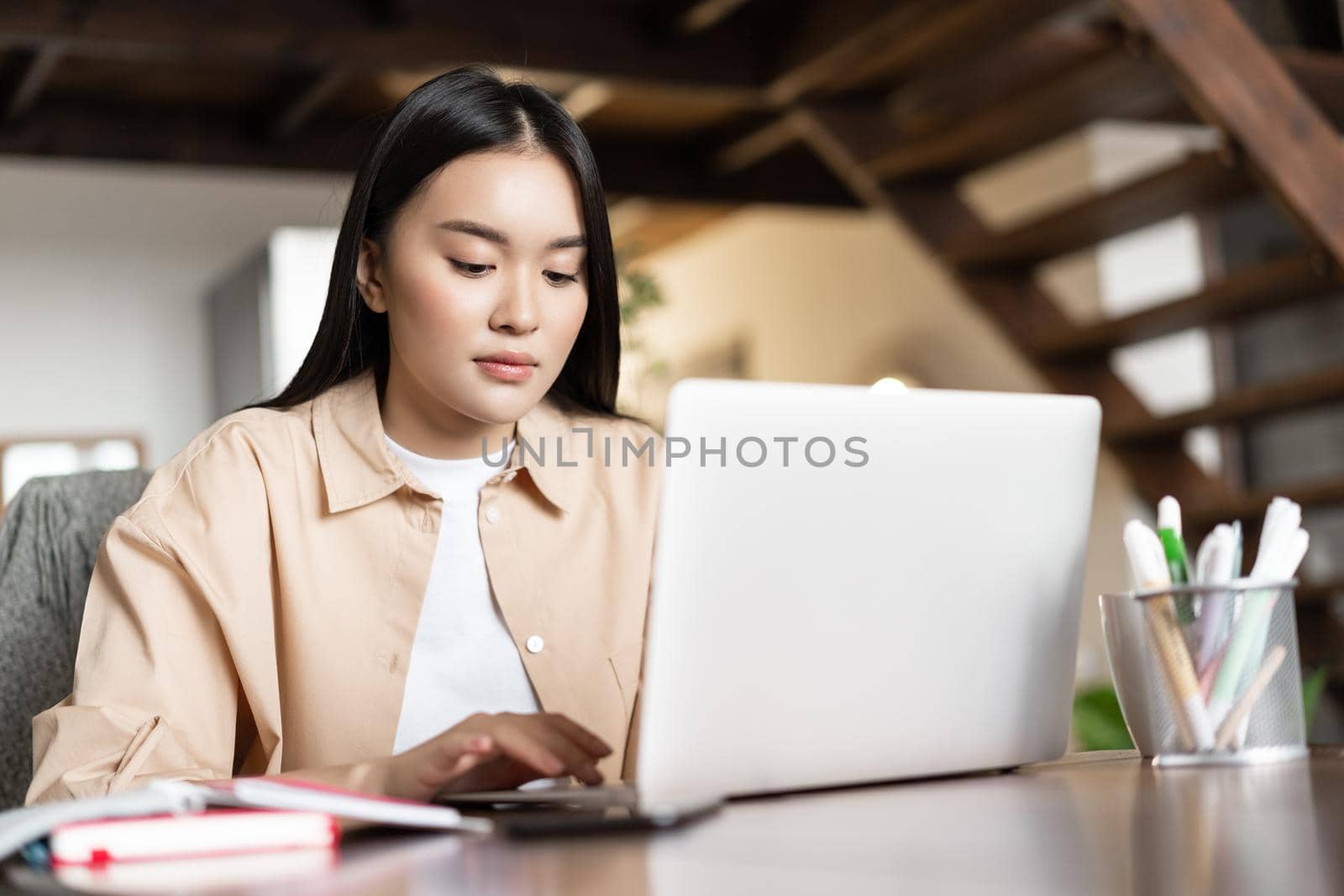 Busy young asian woman working from home, using laptop, sitting at table with books and pencils, typing on computer keyboard.