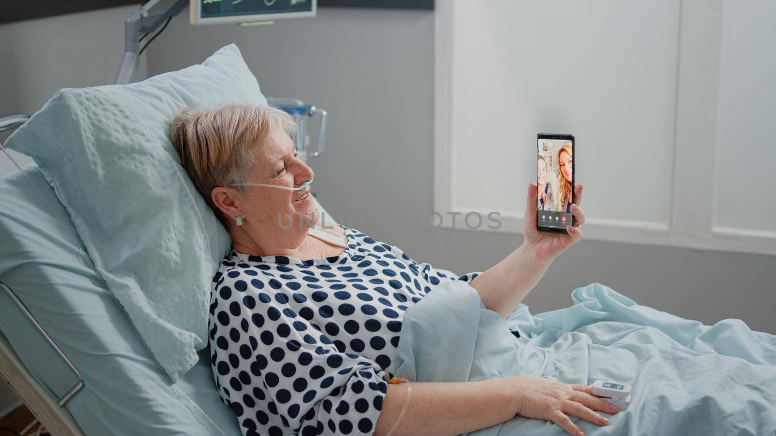 Senior patient talking to family on video call in hospital ward bed. Woman with sickness using smartphone and online video conference for remote communication with niece and daughter.
