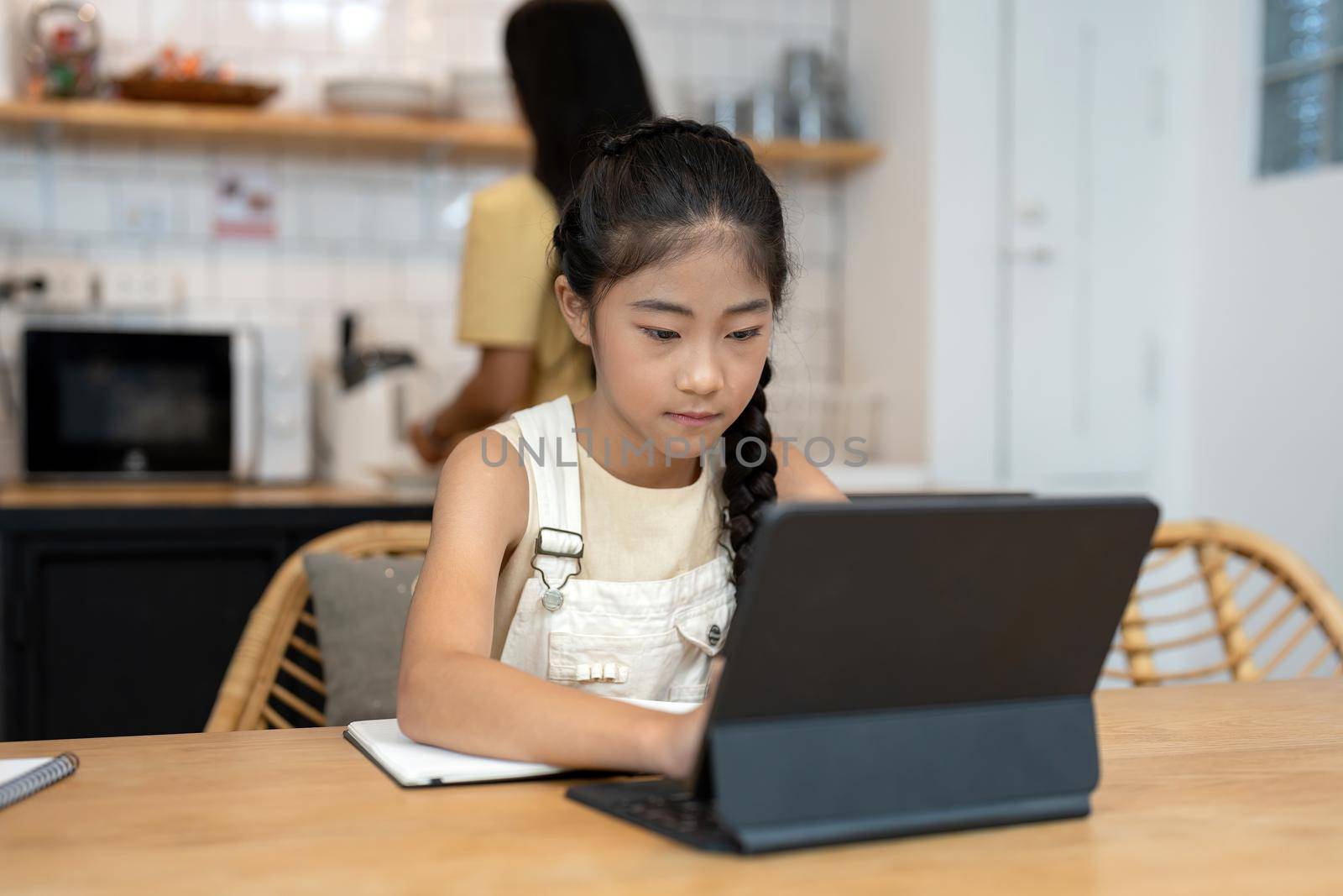 Portrait of little girl using laptop during online class with tutor or teacher while sitting at desk in cozy kitchen interior