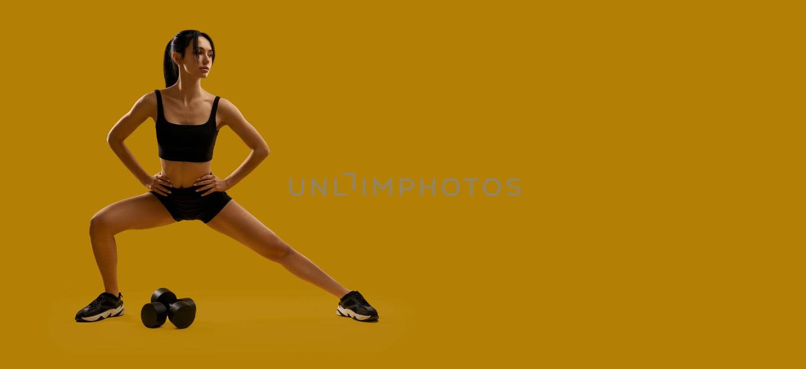 Front view of sporty, slim female exercising, working out, training. Young woman stretching, making step with hands on waist, looking aside. Isolated on yellow studio background.