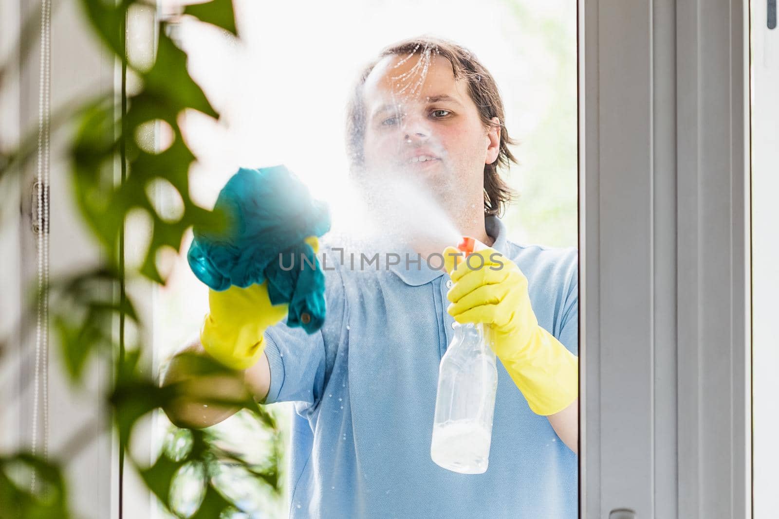 Husband wearing yellow rubber gloves cleaning windows with blue rag at home. Selective focus on the face
