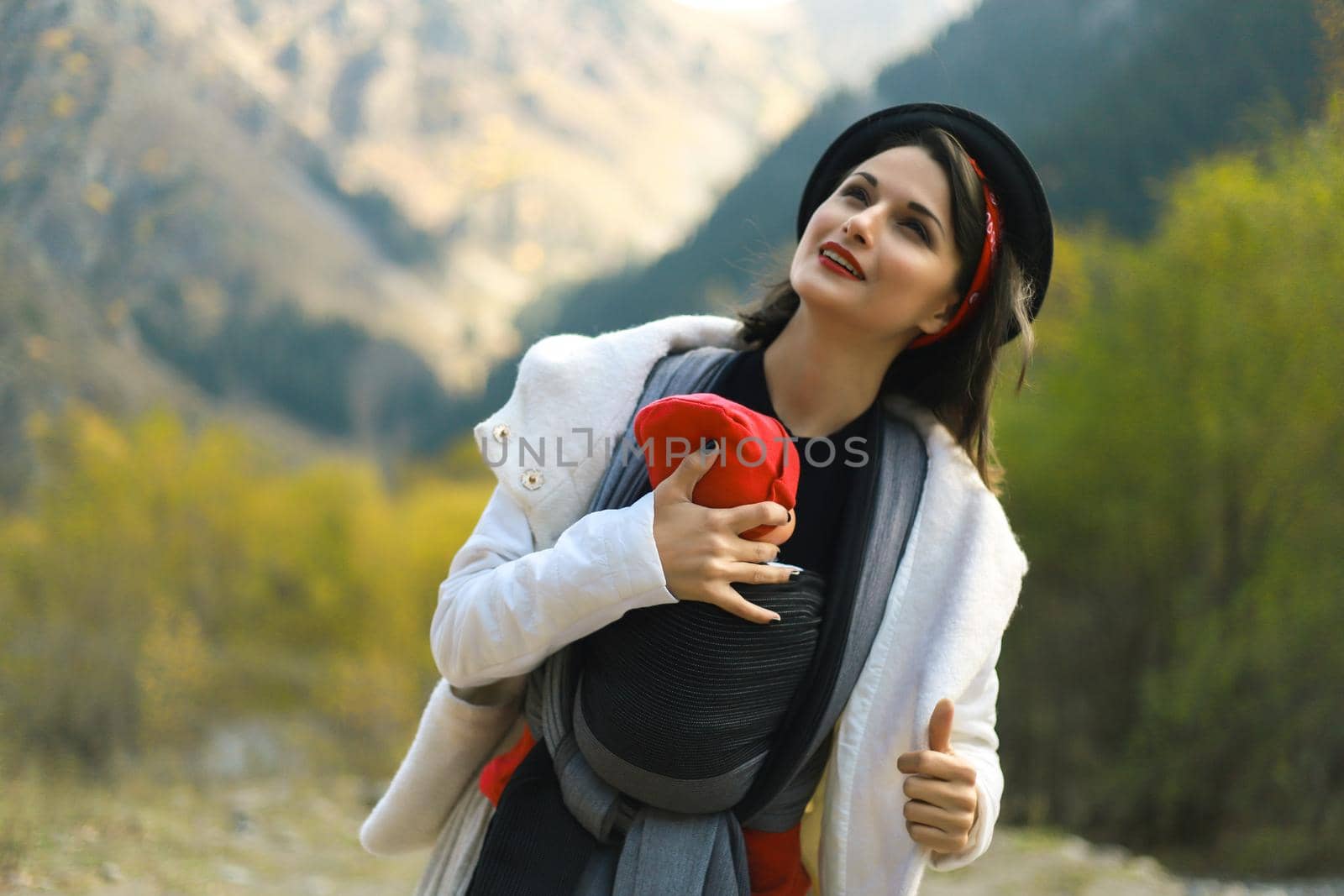 Beautiful young mother with her infant baby in sling outdoor. Woman is carrying her child and travel in autumn mountain. Babywearing concept by Rom4ek