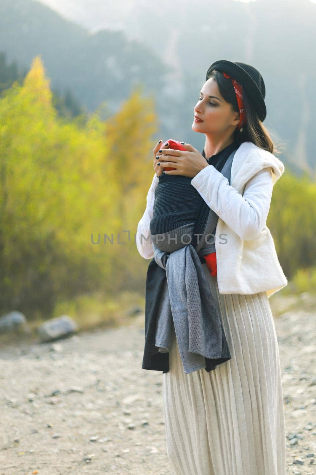 Beautiful young mother with her infant baby in sling outdoor. Woman is carrying her child and travel in autumn mountain. Babywearing concept by Rom4ek