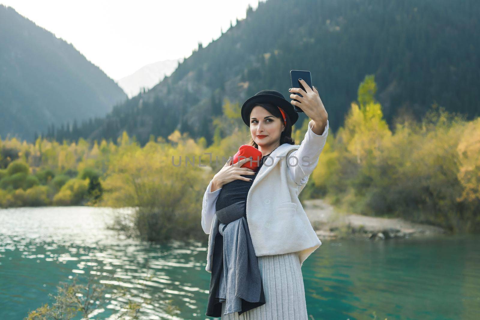 Beautiful young mother with her infant baby in sling make selfie. Mother is carrying her child and travel in autumn mountain lake. Active mother concept