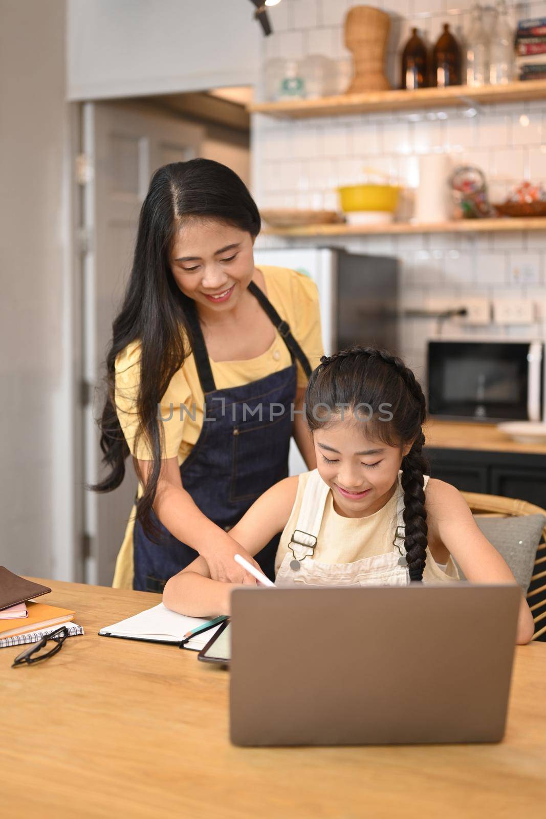 Caring asian mother helping daughter doing homework in kitchen table.