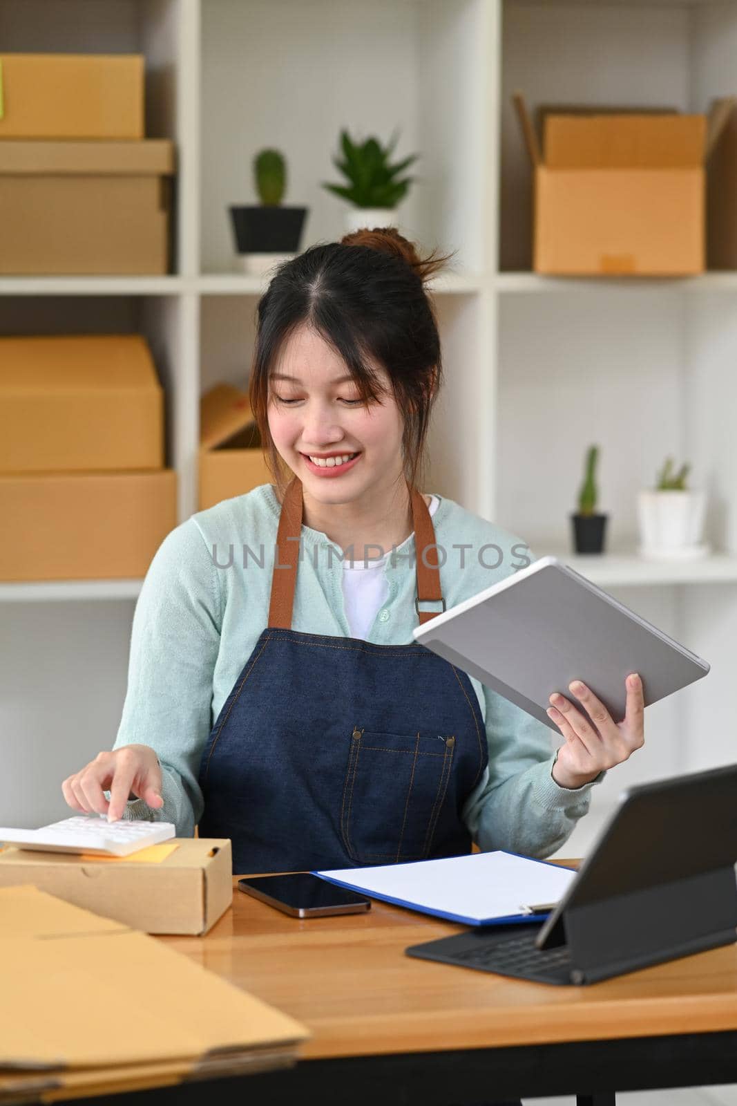 Asian woman small business owner confirming orders or taking orders from customer on digital tablet and using calculator.