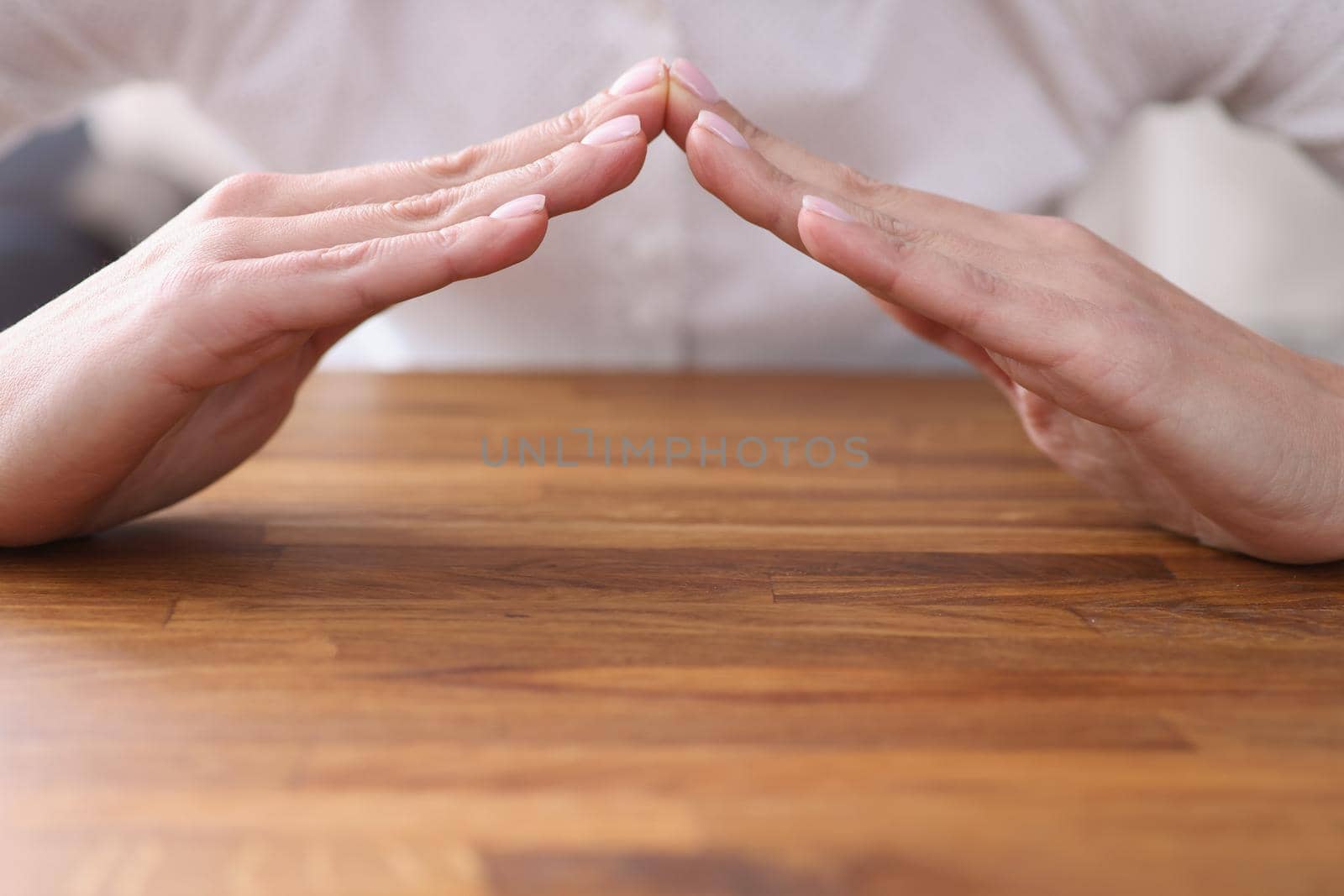 Close-up of female sitting at wooden table and forming hands house roof symbol with hands. Home safety and property security. Real estate insurance concept