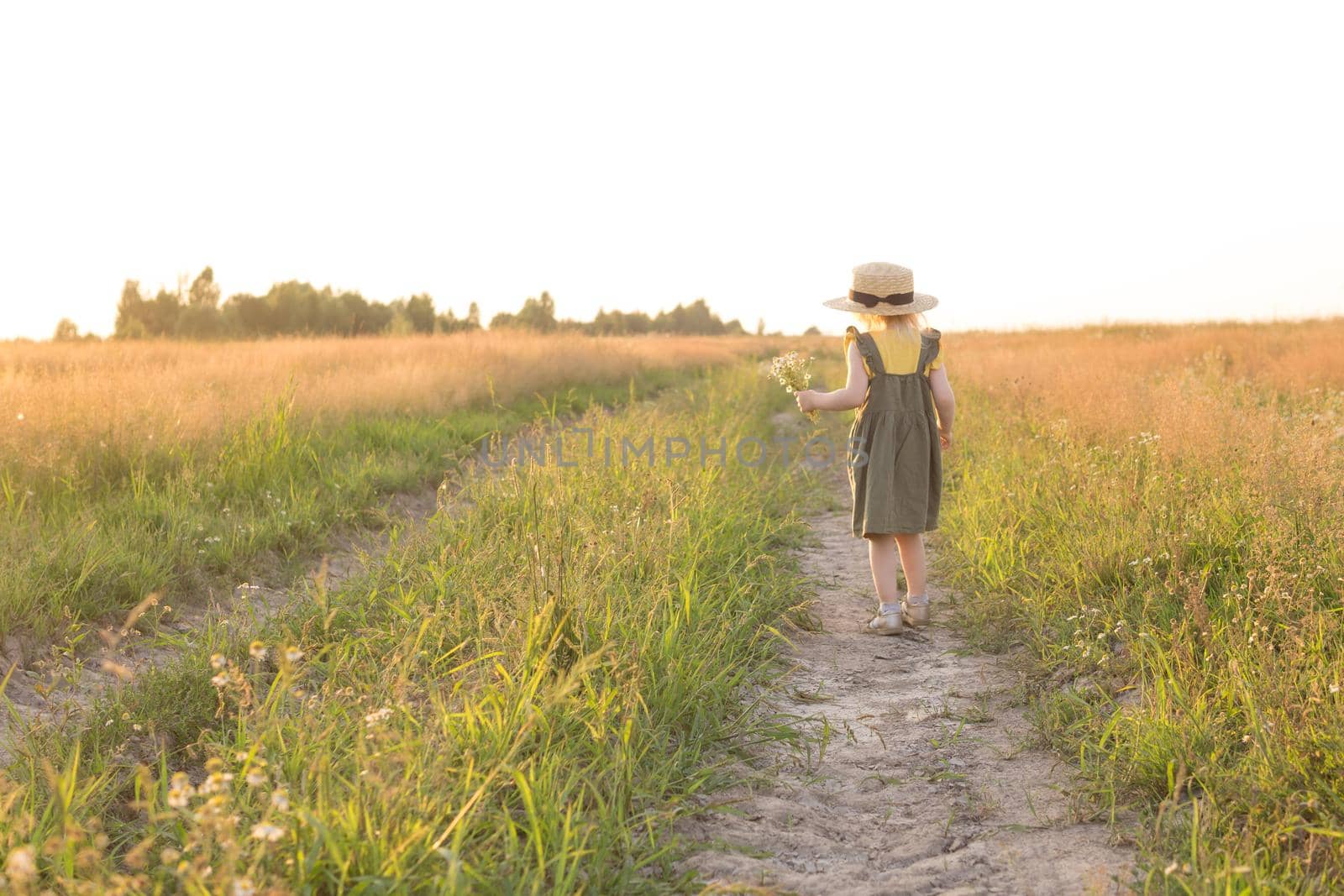 A little blonde girl in a straw hat walks in a field with a bouquet of daisies. The concept of walking in nature, freedom and an eco-friendly lifestyle.