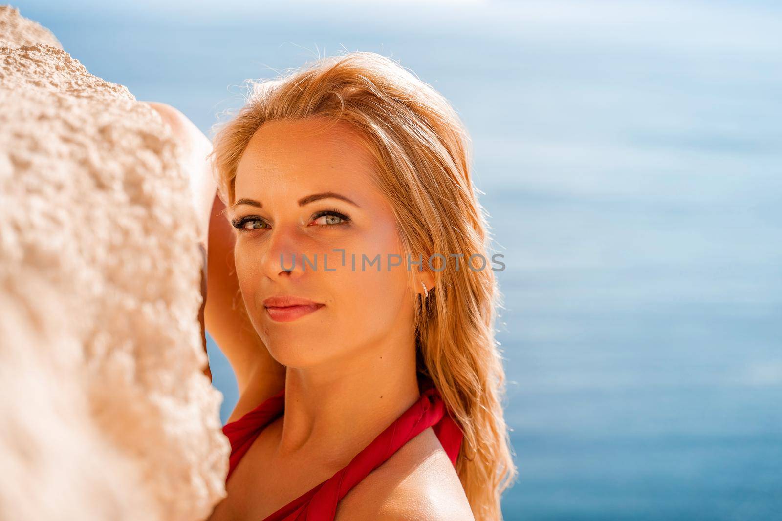 Smiling young woman in a red dress looks at the camera. A beautiful tanned girl enjoys her summer holidays at the sea. Portrait of a stylish carefree woman laughing at the ocean