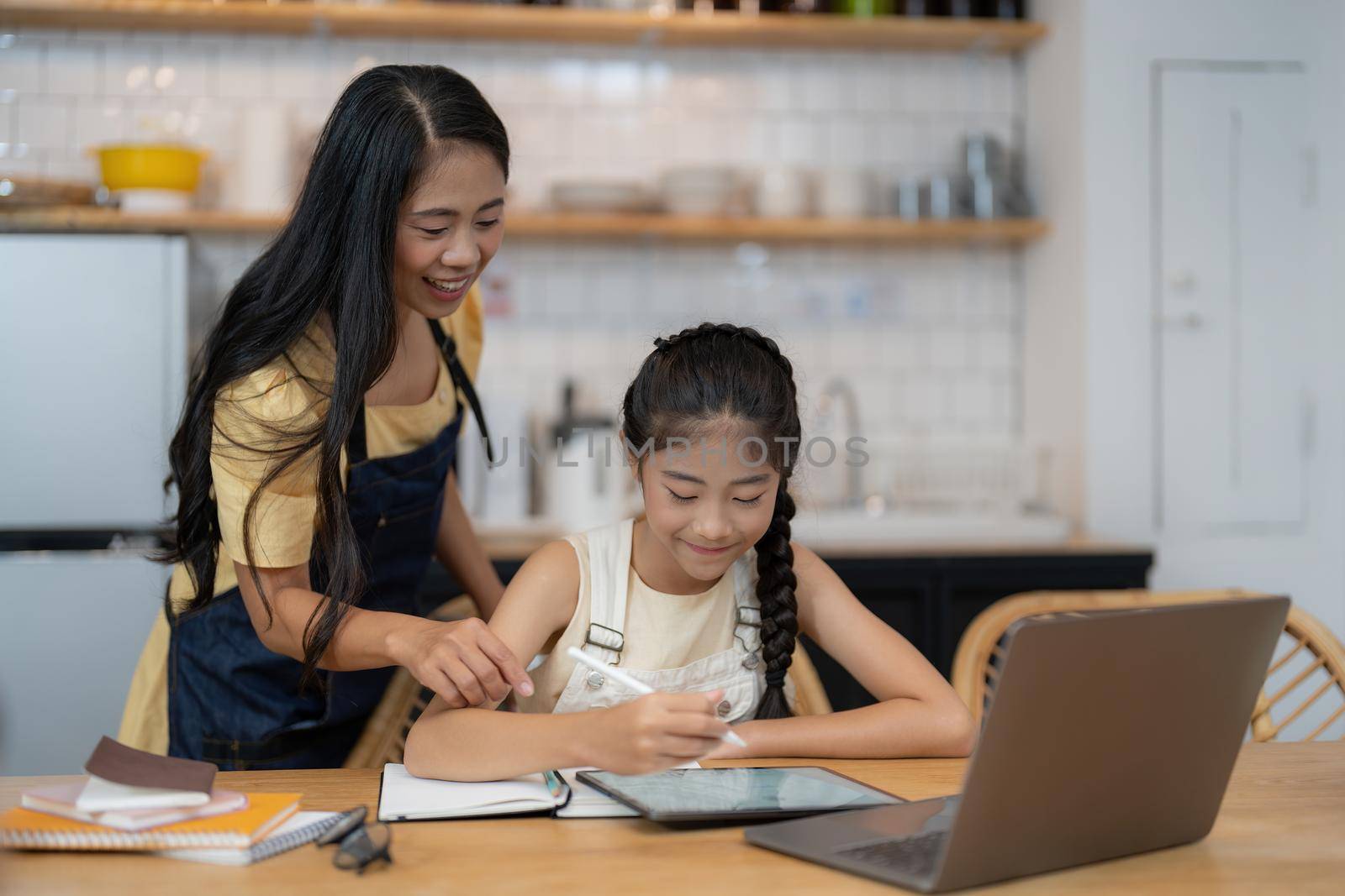 Asian mother and little daughter sit at desk in kitchen studying online together, biracial mom and small girl child handwrite, do homework, learning at home, homeschooling concept by nateemee