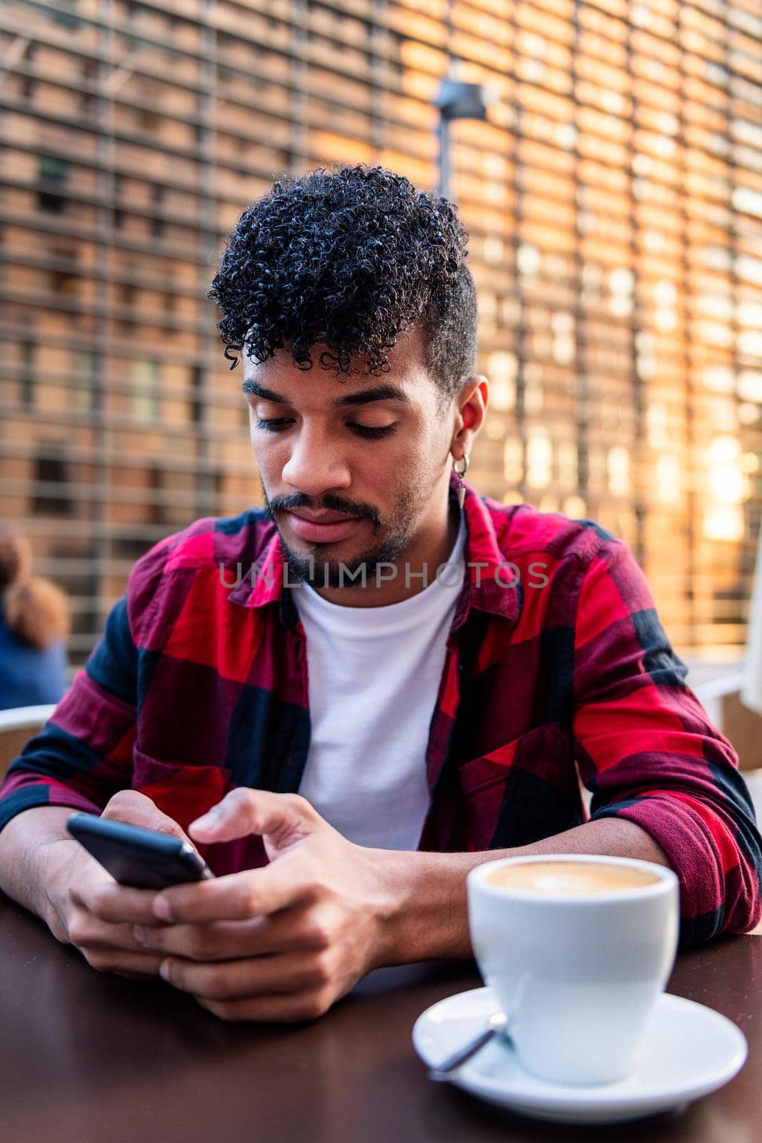vertical photo of a young latin man sitting on a terrace with a coffee and looking at his phone, concept of technology and urban lifestyle