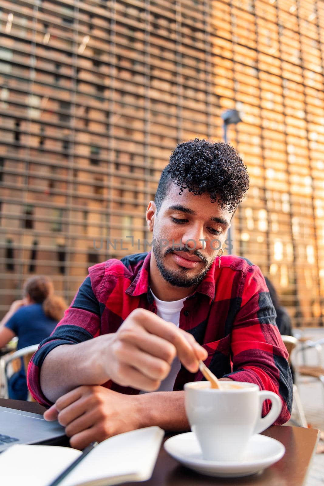 vertical photo of a young latin man stirring his coffee with a spoon while working on the table of the terrace of a coffee shop, concept of urban lifestyle