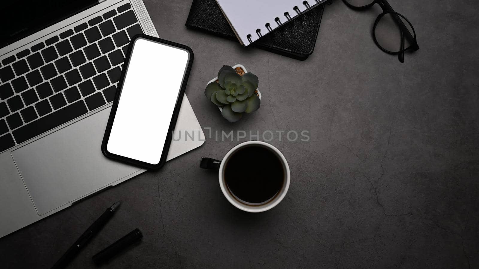 Flat lay smart phone, laptop computer, coffee cup, notepad and glasses on dark background.