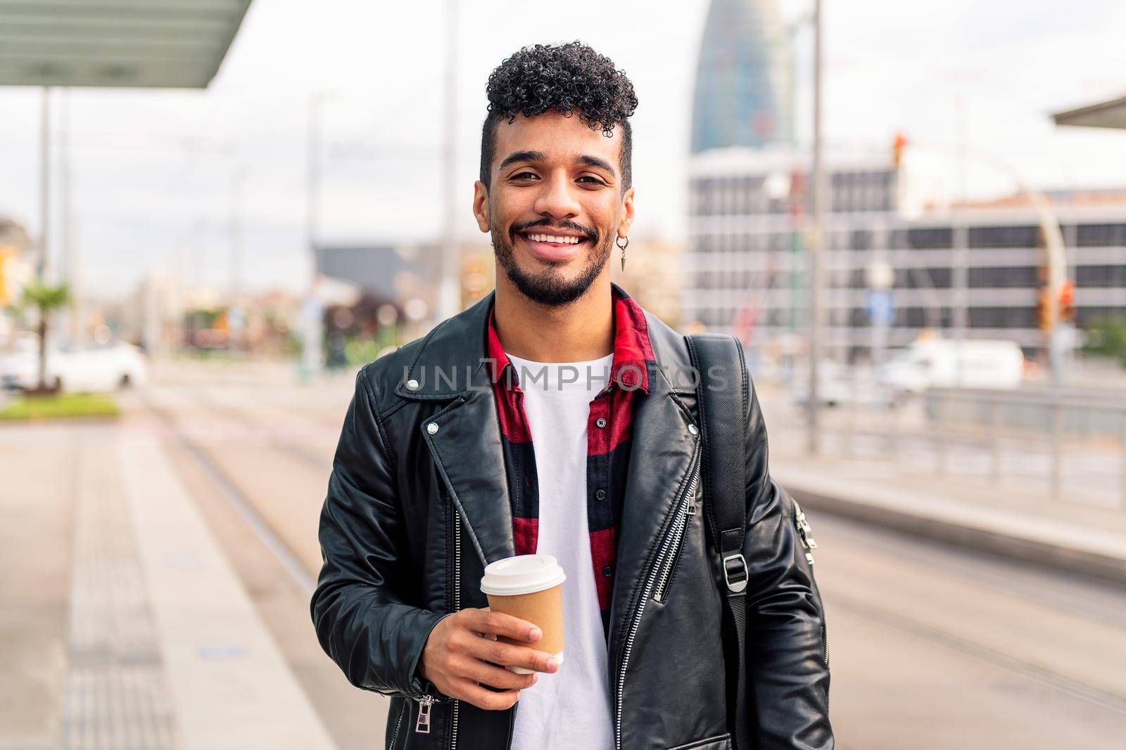 stylish young latin guy smiles happily looking at camera with a take away coffee in his hand, concept of urban lifestyle