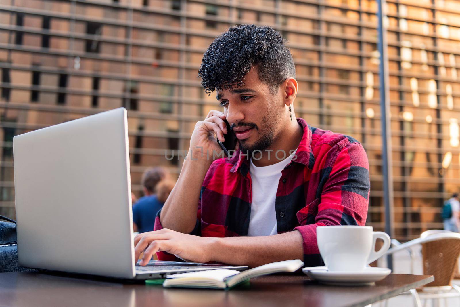 young latin man talking on the phone and typing on his computer on the terrace of a coffee shop in the city, concept of technology and urban lifestyle