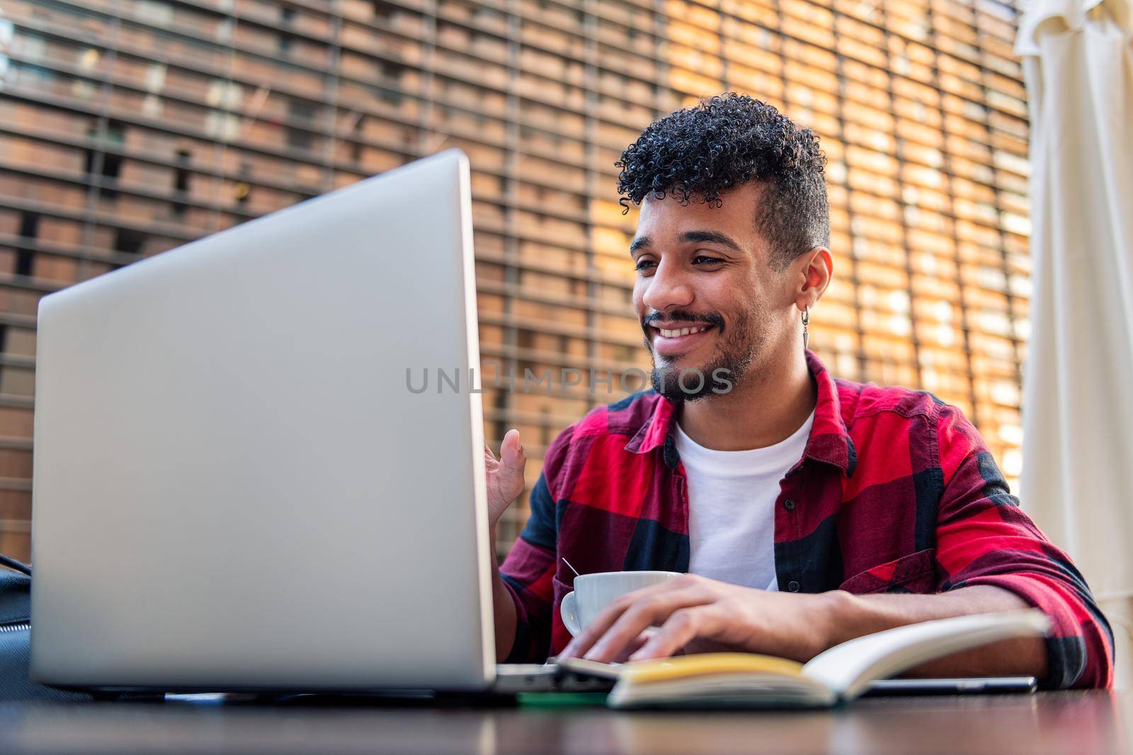 young latin man smiles making a video call with his laptop from the terrace of a coffee shop, concept of technology and urban lifestyle
