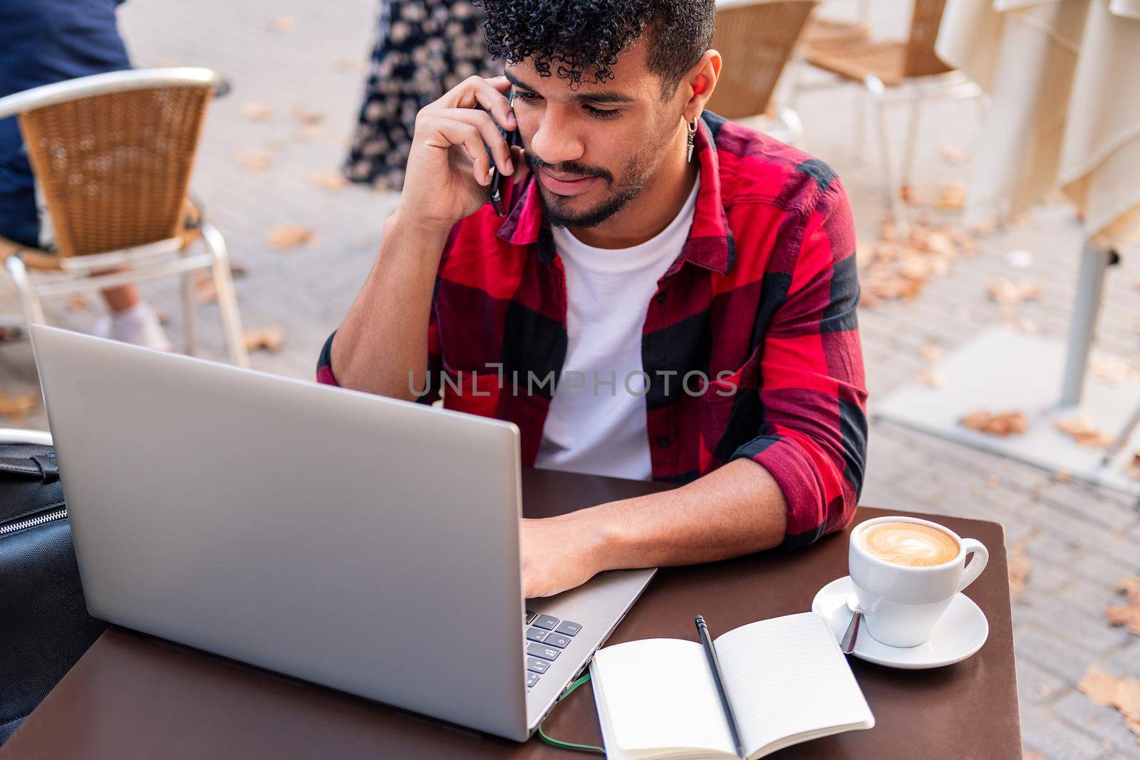 young latin student talking on the phone and typing on his computer on the terrace of a coffee shop in town, concept of technology and urban lifestyle