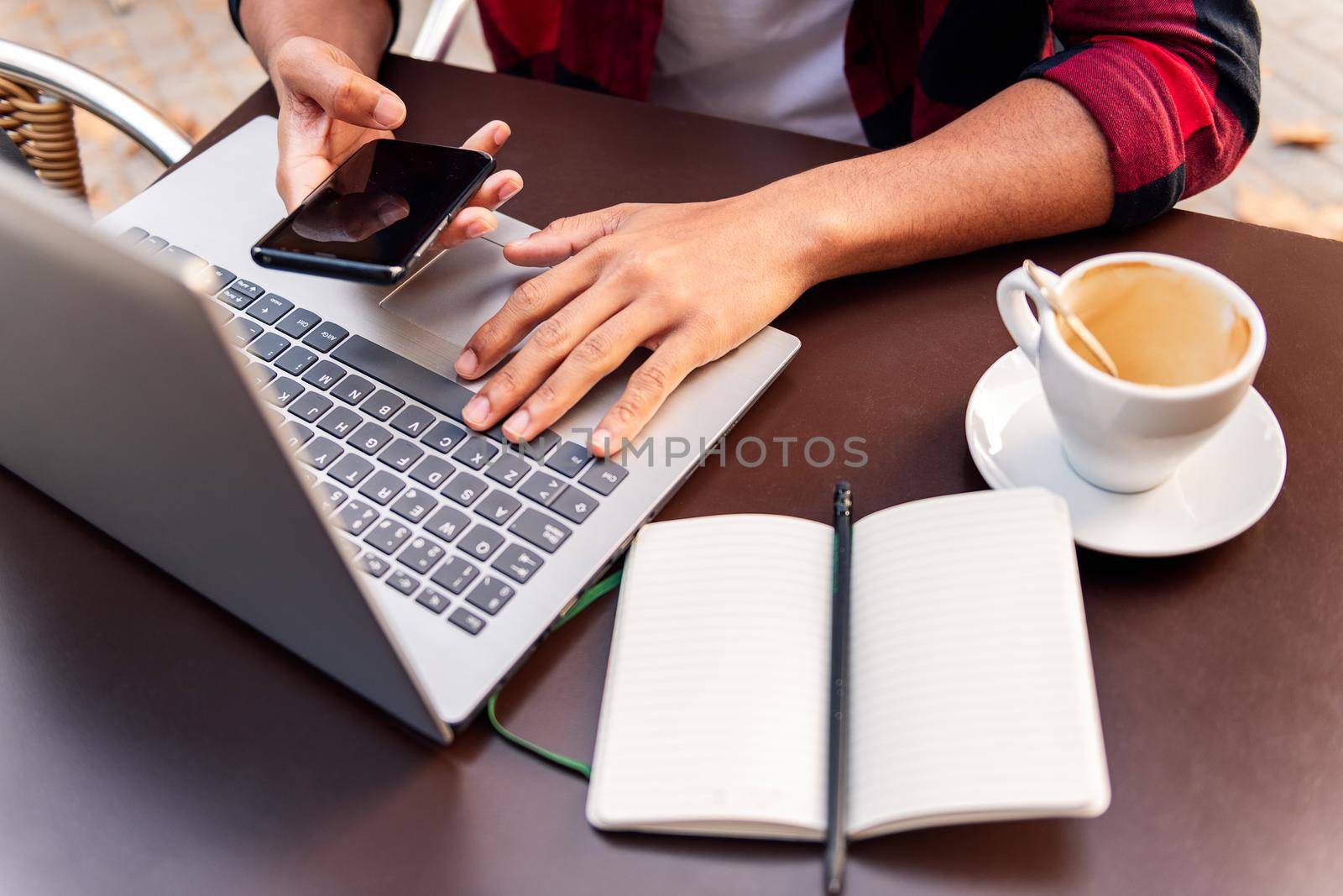 hands of an unrecognizable man working with his computer and phone at the table of the terrace of a coffee shop, concept of technology and urban lifestyle
