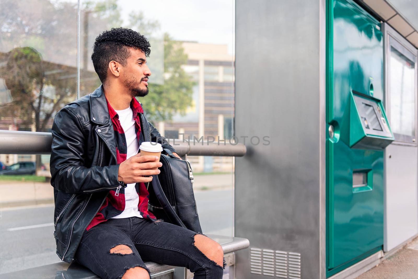 young latin man with a coffee in his hand waits for the arrival of public transportation sitting at the bus stop, concept of sustainable transportation and urban lifestyle