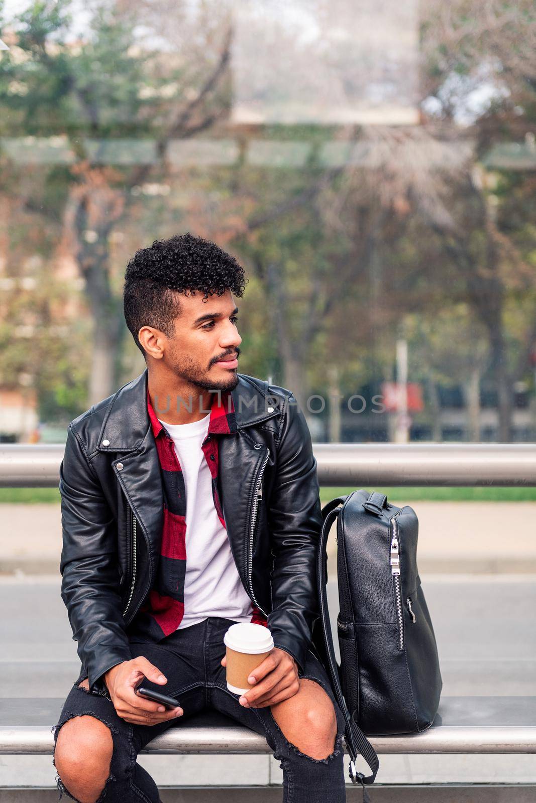stylish latin student with phone and coffee in his hand waits for the arrival of public transportation seated at the bus stop, concept of sustainable transportation and urban lifestyle