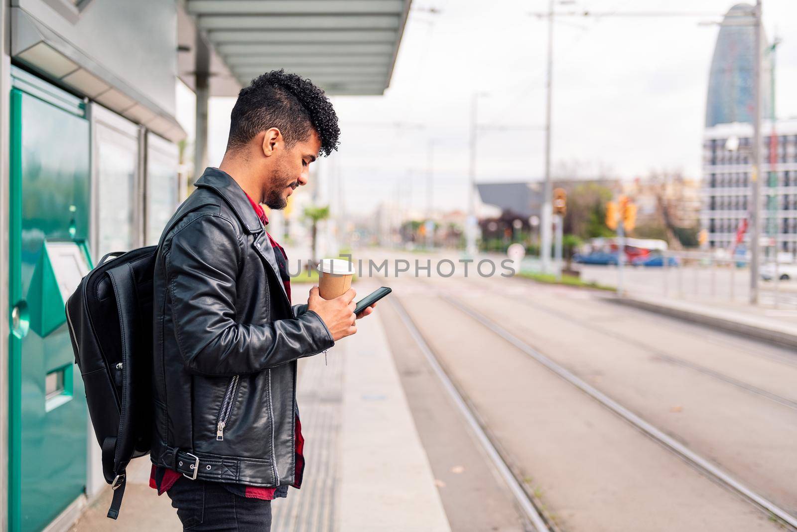 stylish latin student with coffee in hand consults his phone while waiting for public transportation to arrive at the streetcar stop, concept of sustainable transportation and urban lifestyle