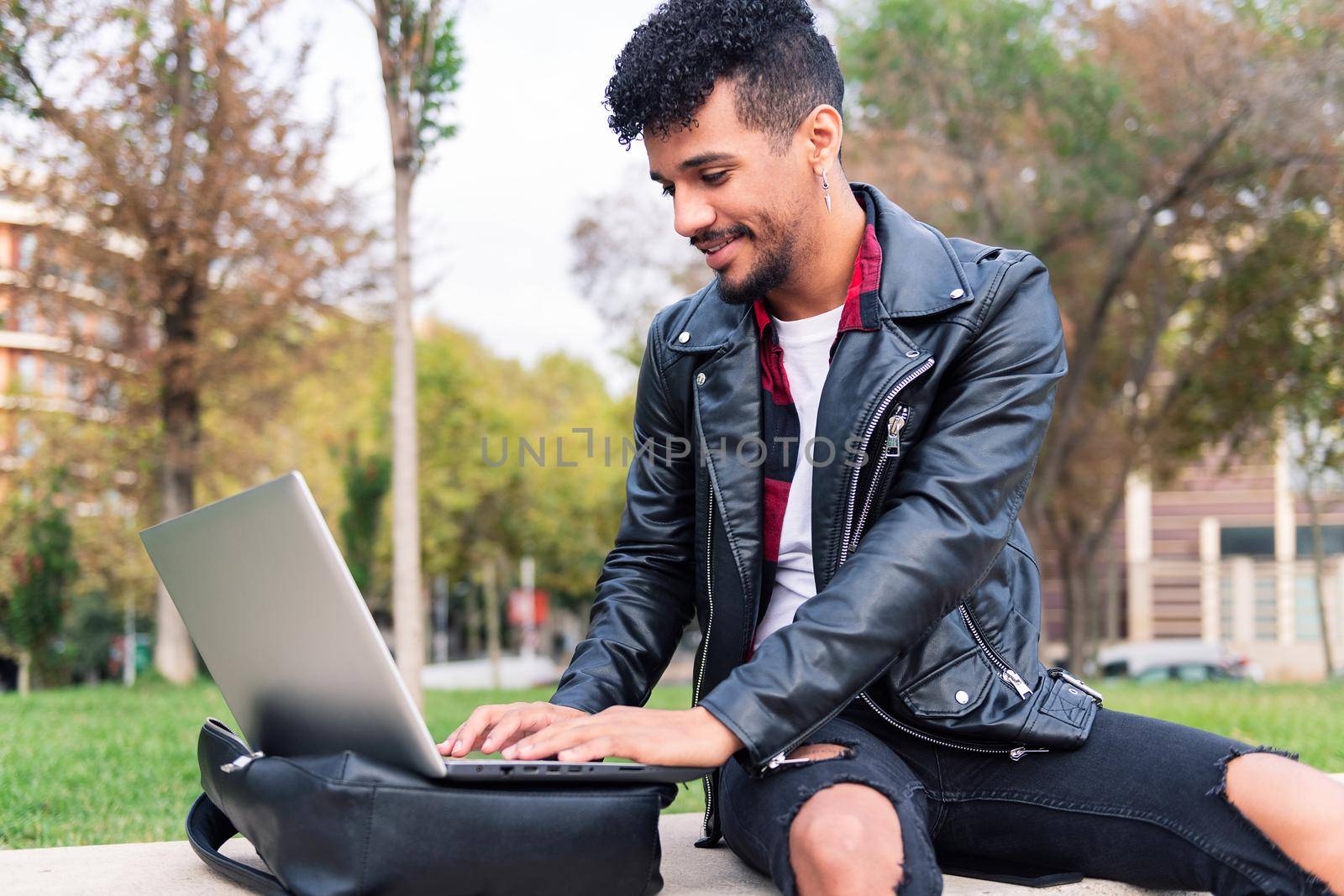 young latin man typing on his laptop computer sitting on a bench in a city park, concept of technology and urban lifestyle
