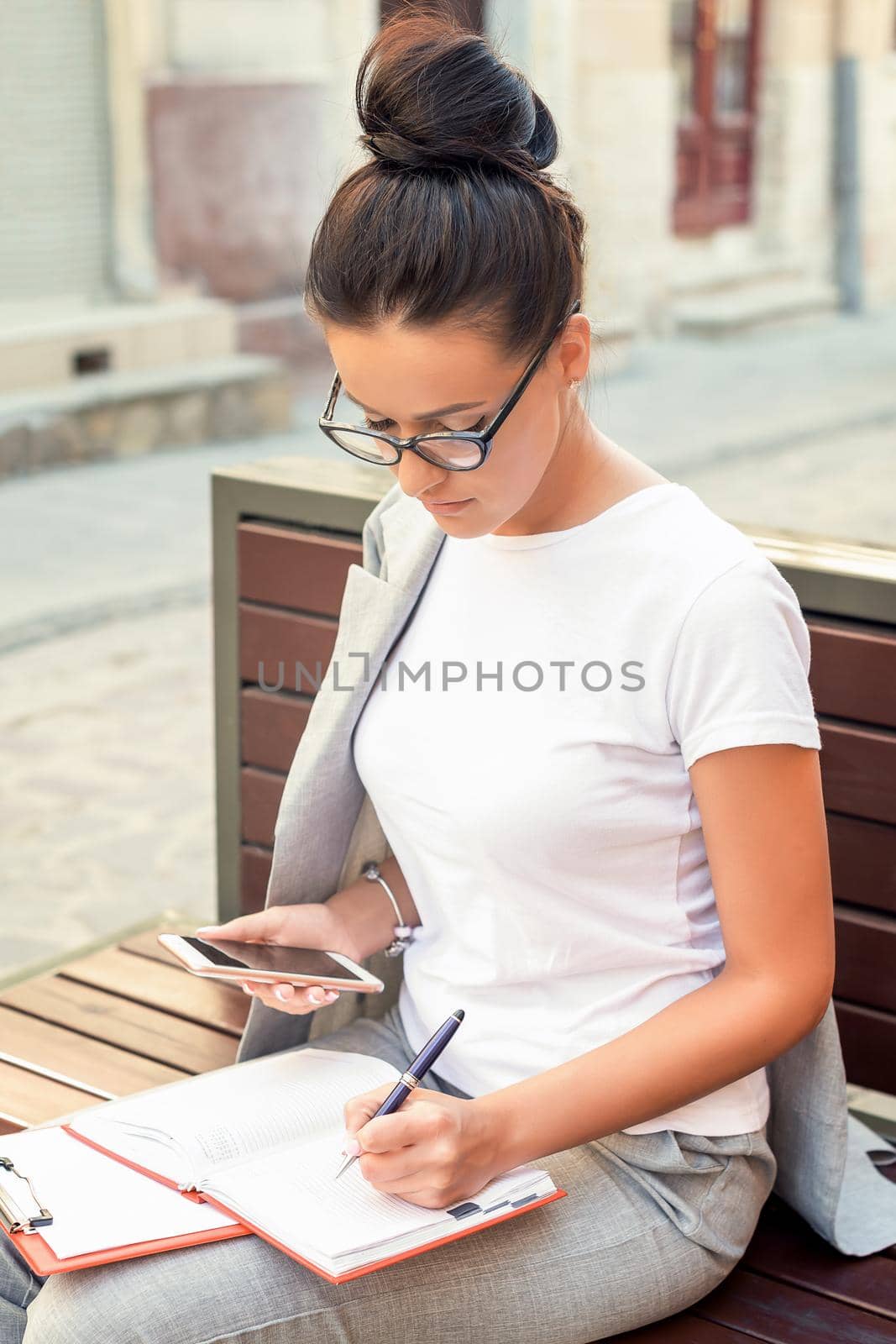 Beautiful young woman is making notes in notebook while is sitting on the bench at the street. Lifestyles concept