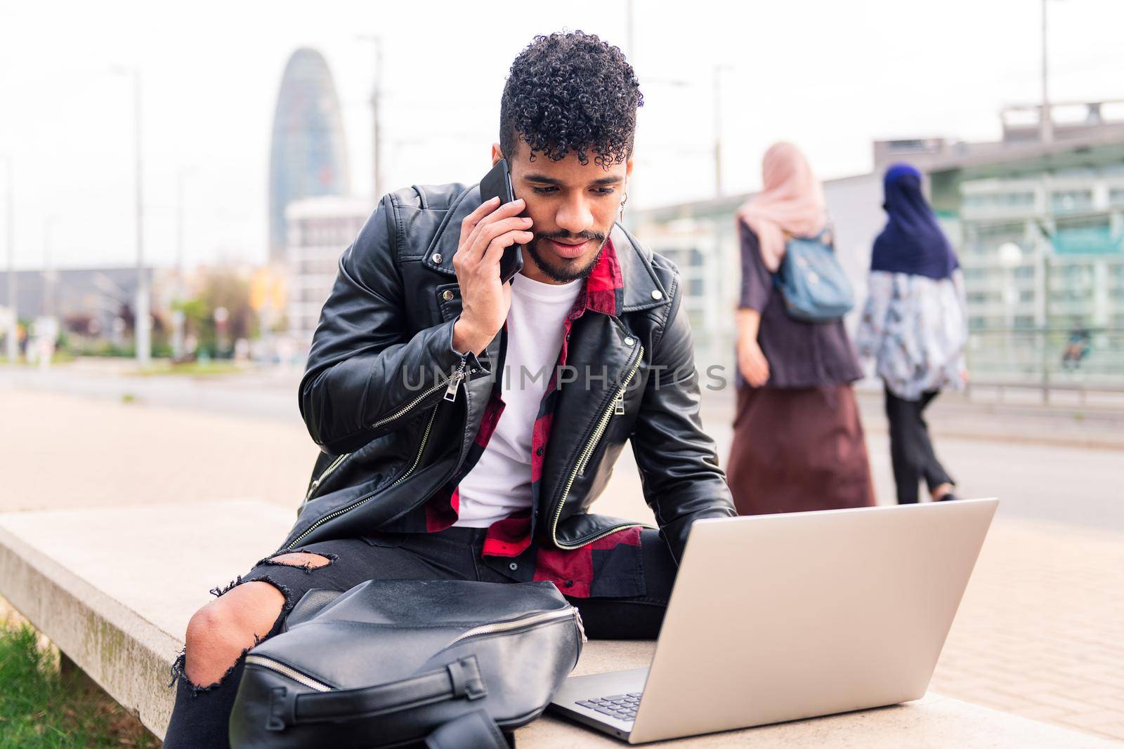 stylish latin man typing on his computer while talking on the phone sitting in the city as two arab women walk past him, concept of technology and urban lifestyle