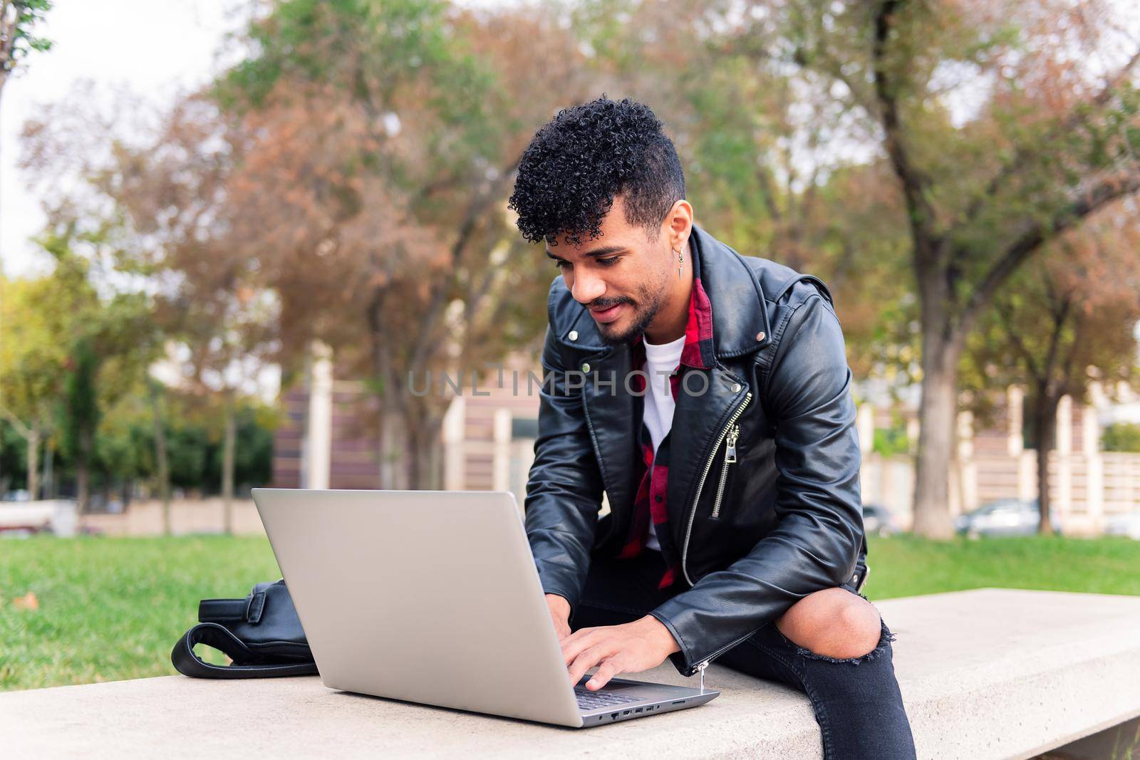 stylish latin student typing on his computer sitting on a park bench in the city, concept of technology and urban lifestyle