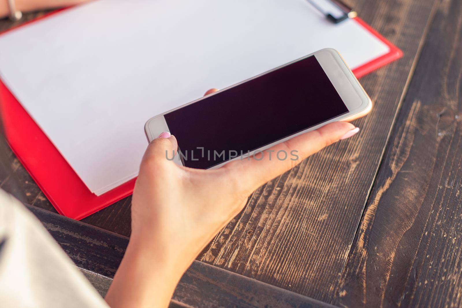 Mockup image of a woman's hands holding white mobile phone with blank black screen on background of white sheet of paper at a cafe.