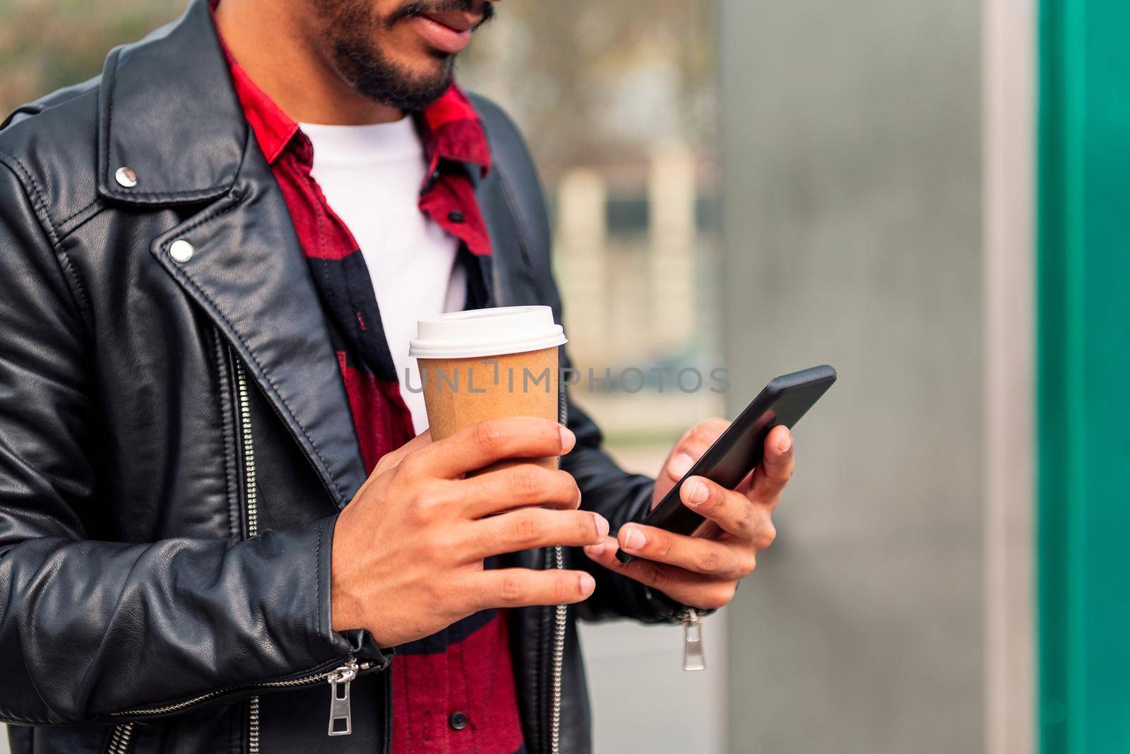 detail of the hands of a modern young latin man consulting his phone with a take away coffee, concept of technology and urban lifestyle