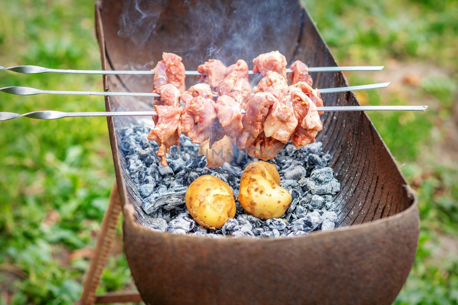 Hands of man prepares barbecue meat with potatoes on skewer by grill on fire outdoors. Concept of lifestyle rustic food preparation
