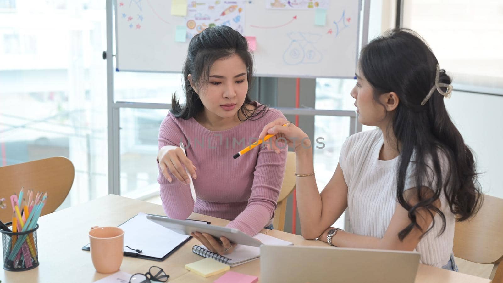 Two young asian women sitting at office desk and talking about project startup ideas. by prathanchorruangsak