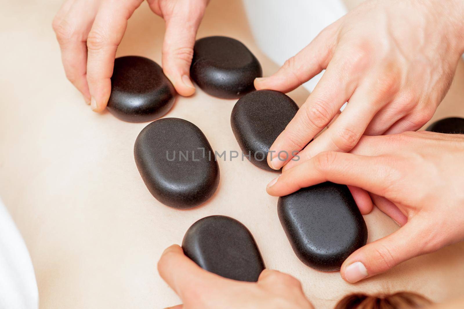 The male four hands laying large new oval black hot stones for back massage on female back in spa salon, close up.