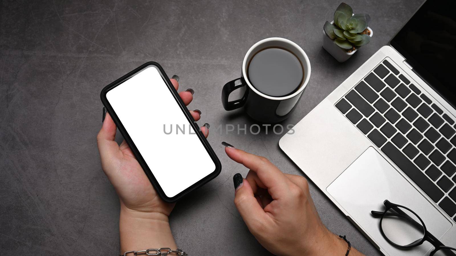 Woman hand holding mobile phone with blank screen on dark stone background. Top view.