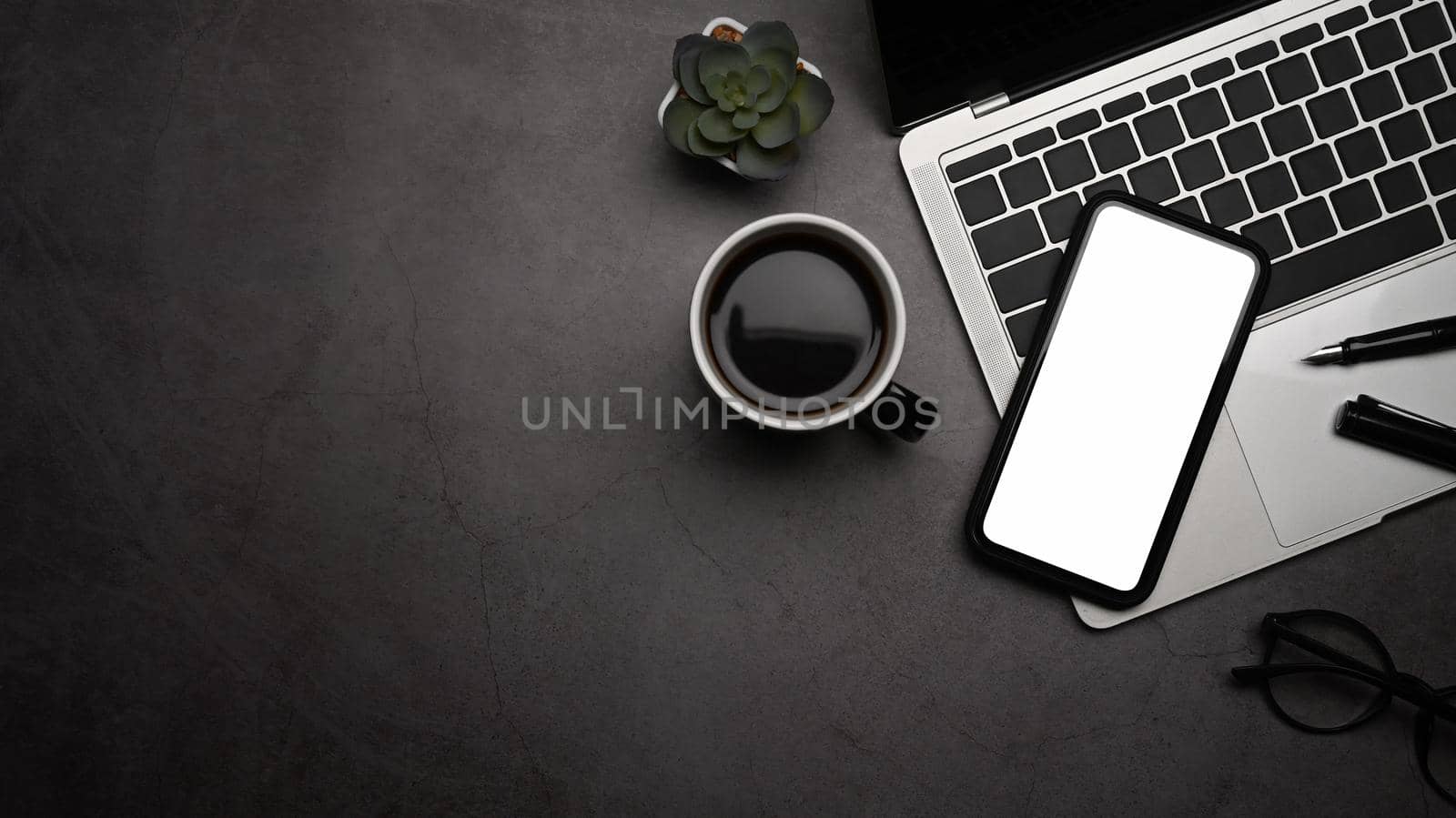 Top view, Flat lay. Smart phone, laptop computer, coffee cup and glasses on dark stone background.