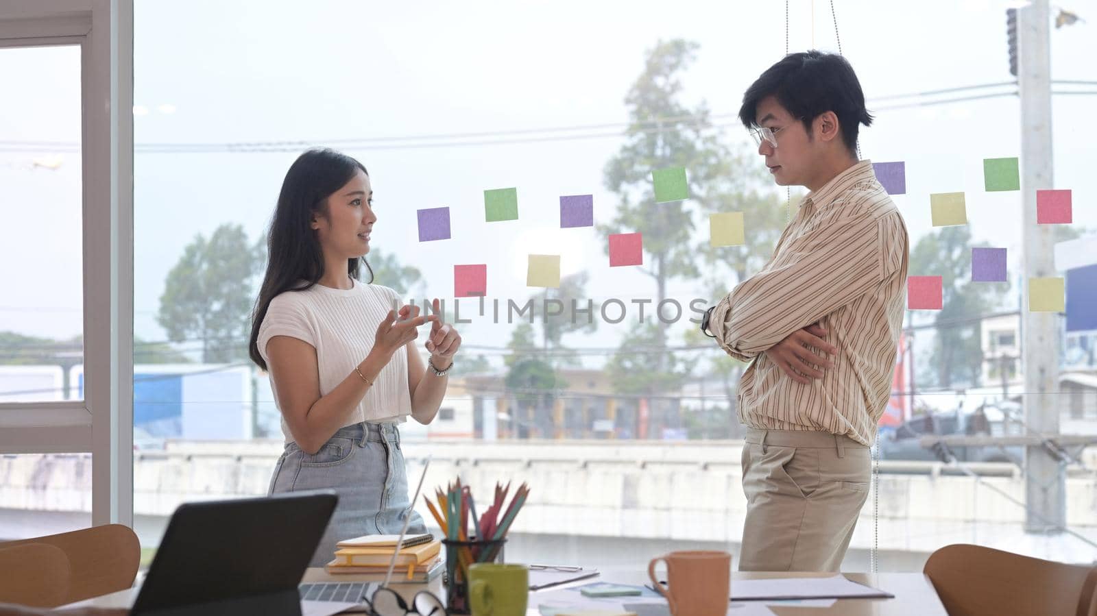 Two young businesspeople standing near large window at office with city buildings in background. by prathanchorruangsak