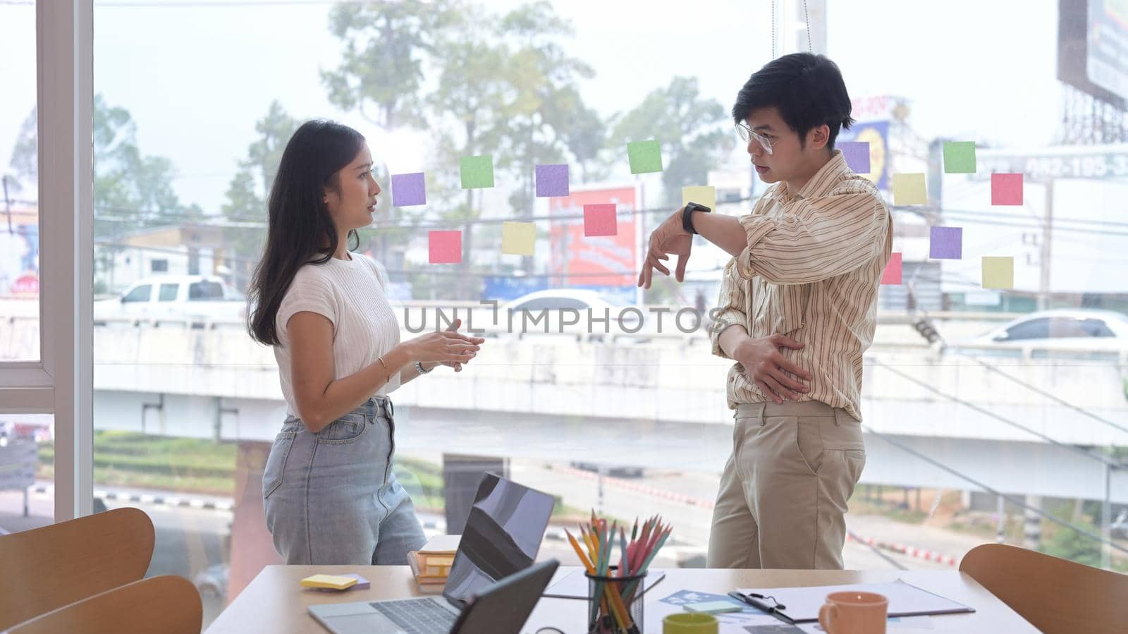 Two young businesspeople standing near large window at office with city buildings in background.