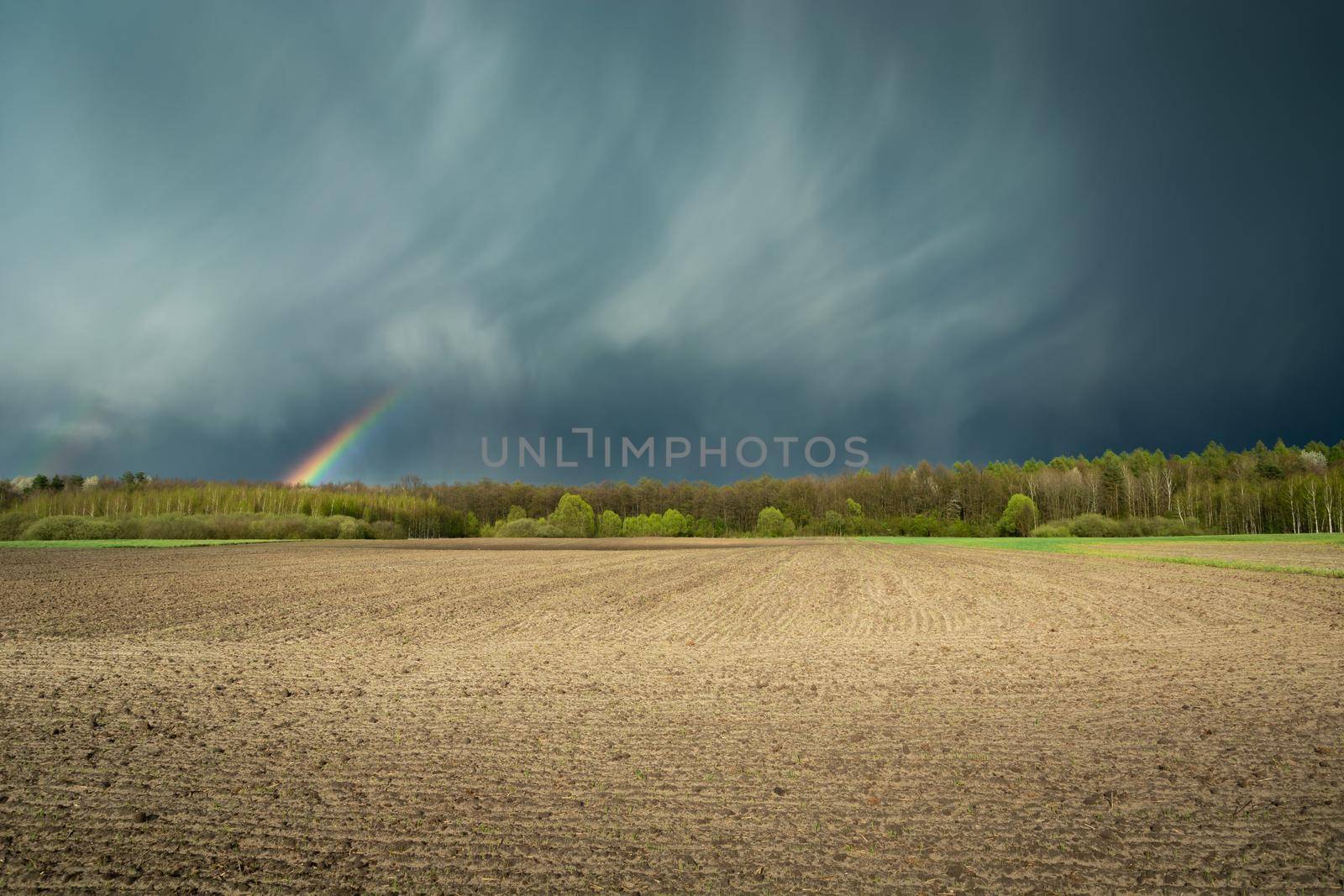 Fantastic clouds with a rainbow and a ploughed field, spring rural view