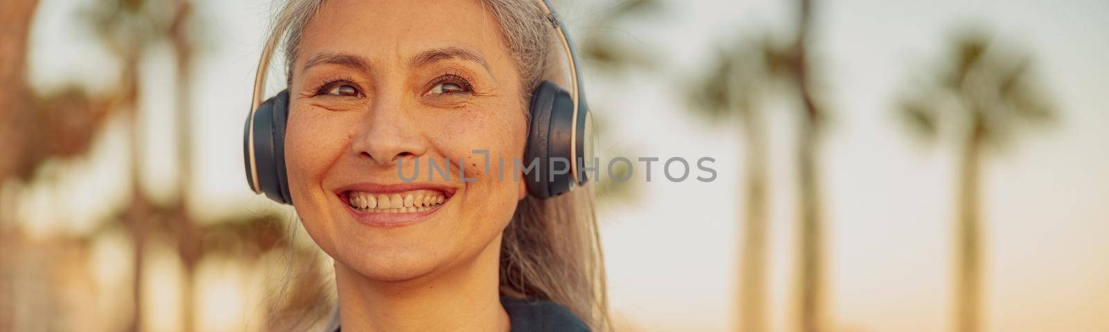 Smiling mature woman listening to music in hedphones standing on background of seafront with palm trees, looking to the side